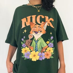 May include: Dark green t-shirt with the word "NICK" in orange above a cartoon fox. The fox is surrounded by flowers and holding a paw-shaped lollipop. The shirt has a loose fit and is made of a soft material.