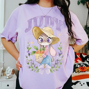 May include: A lilac t-shirt with a cartoon bunny wearing a straw hat and holding carrots. The word "JUDY" is above the character. The design is framed by a floral wreath.