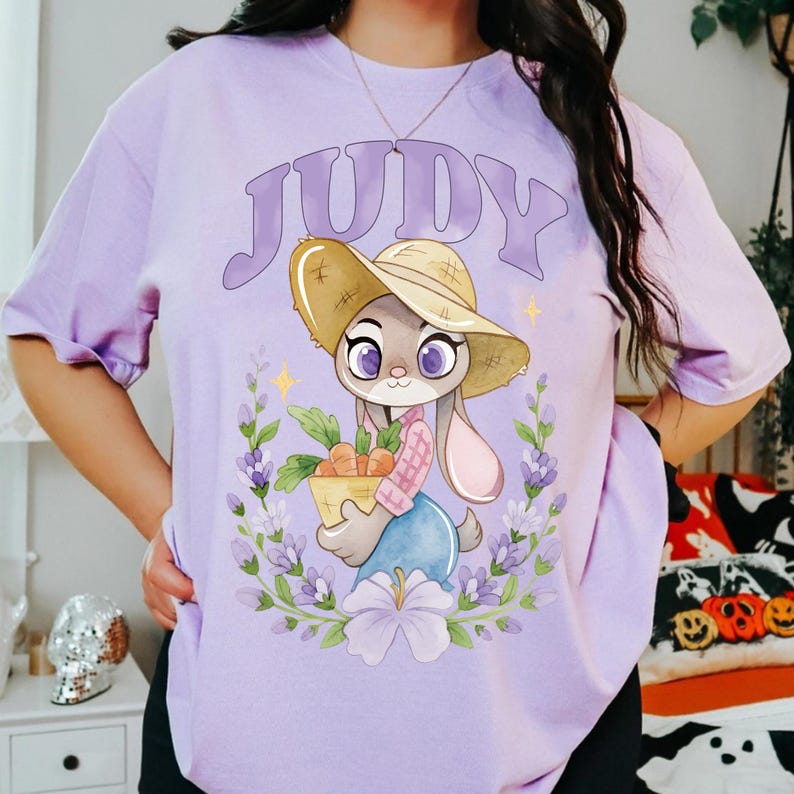 May include: A lilac t-shirt with a cartoon bunny wearing a straw hat and holding carrots. The word "JUDY" is above the character. The design is framed by a floral wreath.