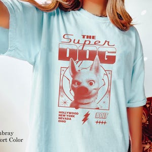 May include: Light blue t-shirt with a red graphic that reads "THE SUPER DOG" above a cartoon dog illustration. Below the illustration are the words "HOLLYWOOD NEW YORK NEVADA OHIO" and a lightning bolt with the word "BOLT".