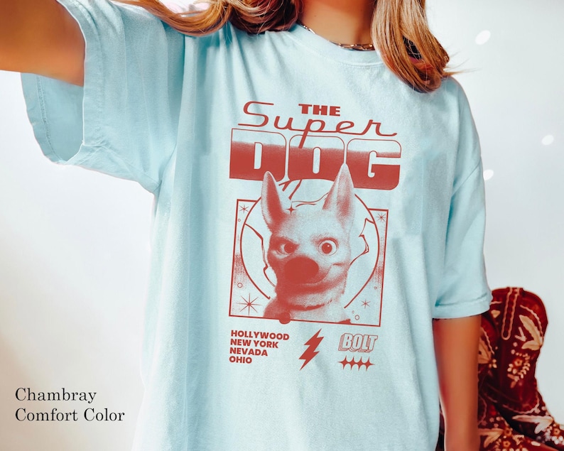 May include: Light blue t-shirt with a red graphic that reads "THE SUPER DOG" above a cartoon dog illustration. Below the illustration are the words "HOLLYWOOD NEW YORK NEVADA OHIO" and a lightning bolt with the word "BOLT".