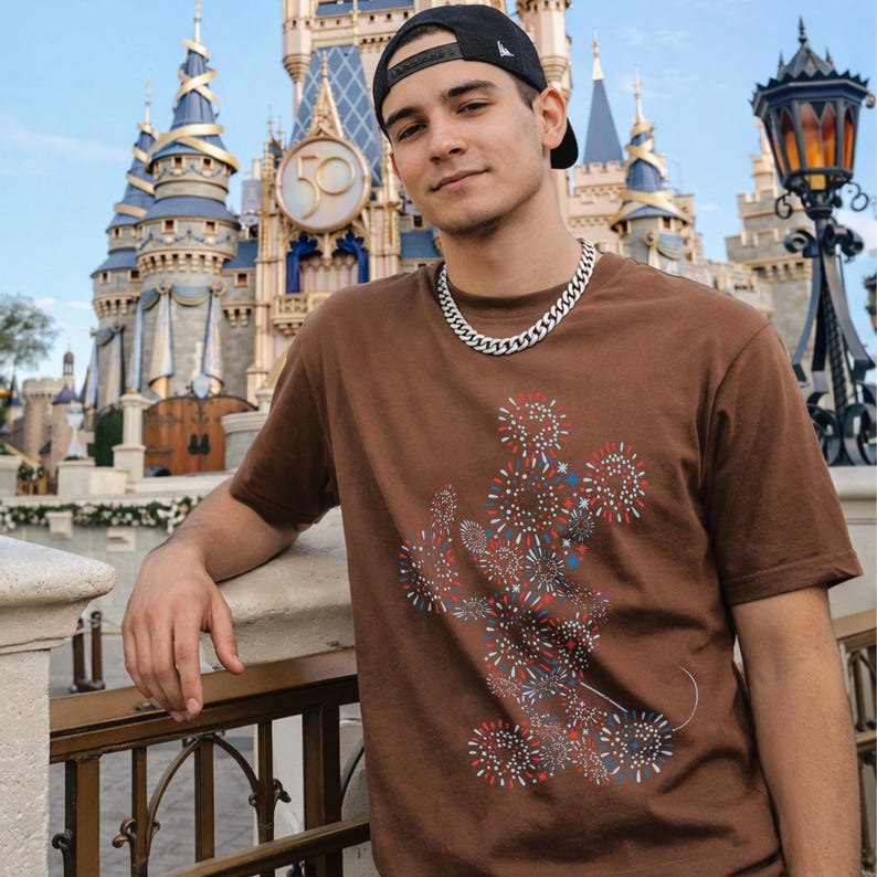 May include: Brown t-shirt featuring a graphic of Mickey Mouse made of fireworks in red, white, and blue. The shirt is paired with a silver chain necklace and a black baseball cap. The background shows a castle.