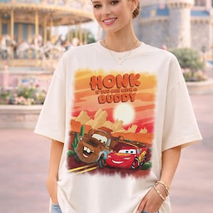 May include: Cream-colored t-shirt featuring a graphic of the Cars characters, Tow Mater and Lightning McQueen, with a desert landscape background. The shirt has the text "HONK IF YOU ARE WITH A BUDDY" in orange and red.