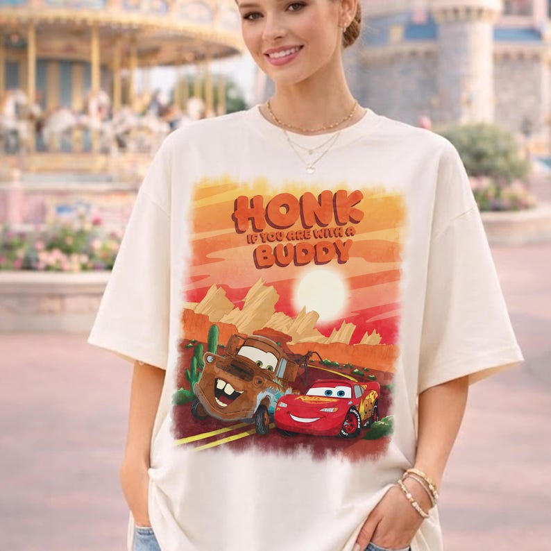 May include: Cream-colored t-shirt featuring a graphic of the Cars characters, Tow Mater and Lightning McQueen, with a desert landscape background. The shirt has the text "HONK IF YOU ARE WITH A BUDDY" in orange and red.