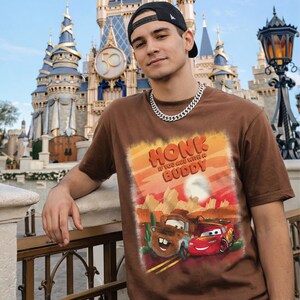 May include: Brown t-shirt featuring a graphic of Mater and Lightning McQueen from the movie Cars, with the text "Honk If You Are With A Buddy". The shirt is worn with a black cap and silver chain.