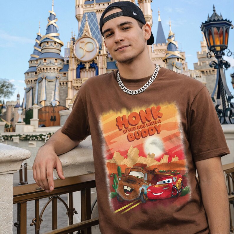 May include: Brown t-shirt featuring a graphic of Mater and Lightning McQueen from the movie Cars, with the text "Honk If You Are With A Buddy". The shirt is worn with a black cap and silver chain.