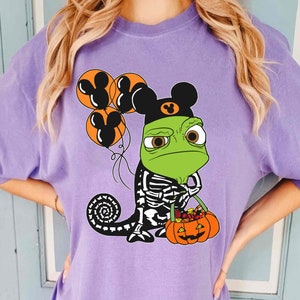 May include: A light purple t-shirt featuring a graphic of a green chameleon wearing a black hat with mouse ears. The chameleon has a skeleton print on its body, holds orange balloons with mouse ear silhouettes, and carries a pumpkin bucket.