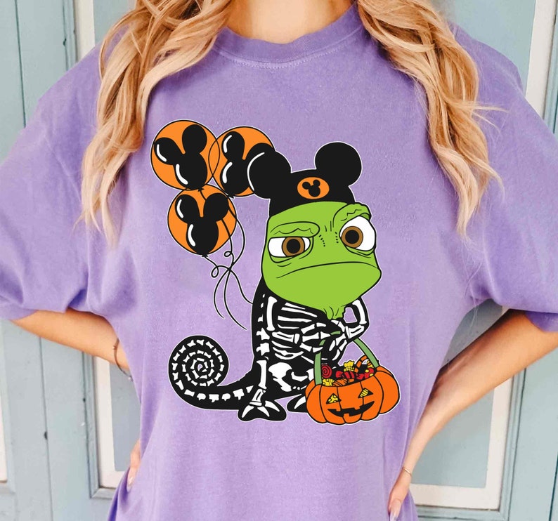 May include: A light purple t-shirt featuring a graphic of a green chameleon wearing a black hat with mouse ears. The chameleon has a skeleton print on its body, holds orange balloons with mouse ear silhouettes, and carries a pumpkin bucket.