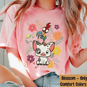 Pu&ograve; includere: Una maglietta rosa con un'immagine di un maiale e un gallo con fiori in stile cartone animato. Il testo "Blossom - Only for Comfort Colors Shirt" &egrave; in basso nell'immagine.