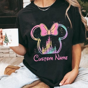 K&ouml;nnte beinhalten: Schwarzes T-Shirt mit einem farbenfrohen Minnie-Mouse-Design, einschlie&szlig;lich Schleife, Schloss und dem Text "Custom Name". Das Design verwendet Pastellfarben und einen Spritzeffekt. Dies ist ein Freizeitkleidungsst&uuml;ck.