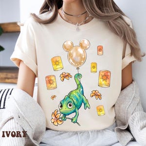 May include: Ivory-colored t-shirt featuring a cartoon chameleon holding a flower and a Mickey Mouse-shaped balloon. The design includes floating lanterns and flowers. The word "IVORY" is printed at the bottom.