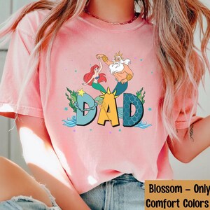 May include: A pale pink t-shirt with a vibrant graphic of Ariel, King Triton, and the word "DAD" in a fun font. The design incorporates aquatic elements such as waves and stars, creating a whimsical theme. The shirt is a Comfort Colors shirt.