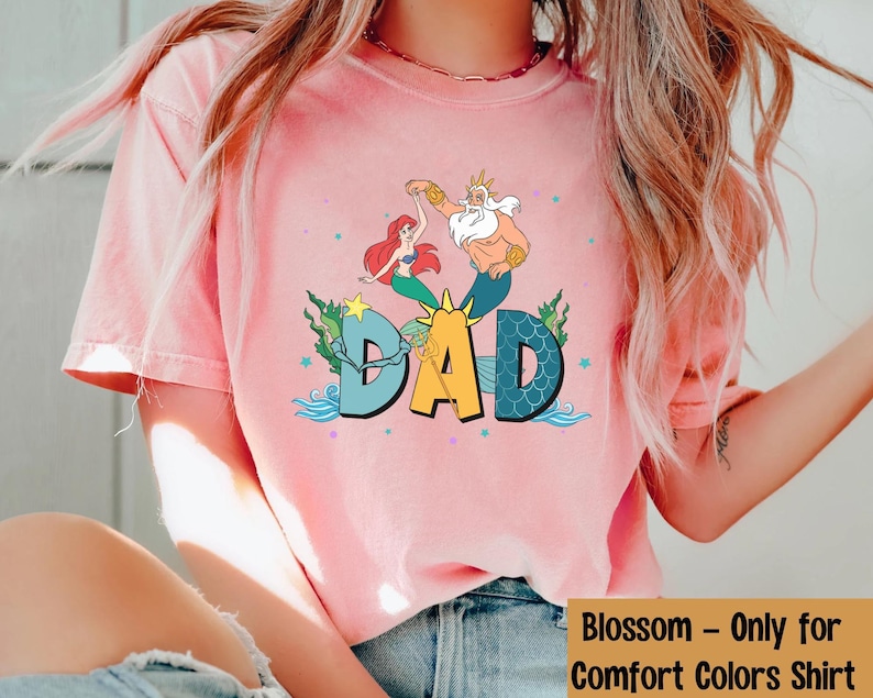 May include: A pale pink t-shirt with a vibrant graphic of Ariel, King Triton, and the word "DAD" in a fun font. The design incorporates aquatic elements such as waves and stars, creating a whimsical theme. The shirt is a Comfort Colors shirt.