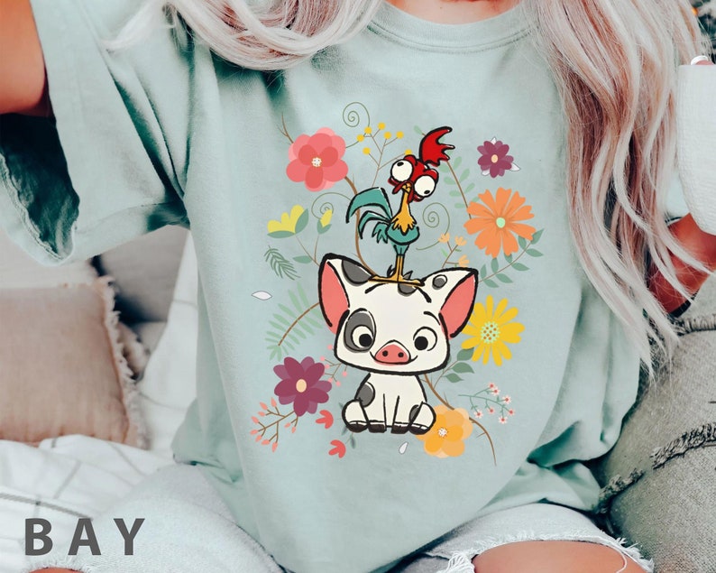 Pu&ograve; includere: Una maglietta blu chiaro con un'immagine di un maiale e un gallo in stile cartone animato con accenti floreali. Il maiale &egrave; bianco con macchie nere e il gallo &egrave; rosso, verde e giallo.