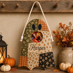 K&ouml;nnte beinhalten: Ein geisterf&ouml;rmiger Wandbehang aus Patchwork-Stoff mit verschiedenen herbstlichen Mustern wie G&auml;nsebl&uuml;mchen, Karos und K&uuml;rbissen. In der Mitte befindet sich eine kreisf&ouml;rmige &Ouml;ffnung, die mit S&uuml;&szlig;igkeiten gef&uuml;llt ist, und der Name Harper steht in schwarzer Schrift.