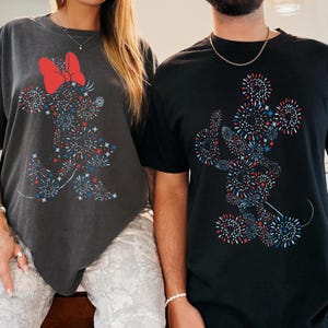 May include: Two t-shirts, one charcoal gray and one black, each featuring a graphic of a character made of red, white, and blue fireworks. The gray shirt has a red bow. The shirts are short-sleeved.