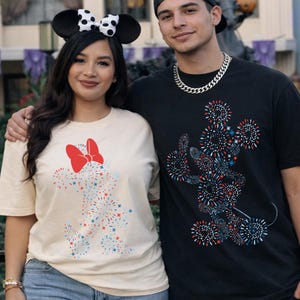May include: Two people wearing matching t-shirts with a Mickey Mouse and Minnie Mouse design made of red, white, and blue fireworks. The woman wears a cream-colored shirt with a red bow and mouse ears. The man wears a black shirt.
