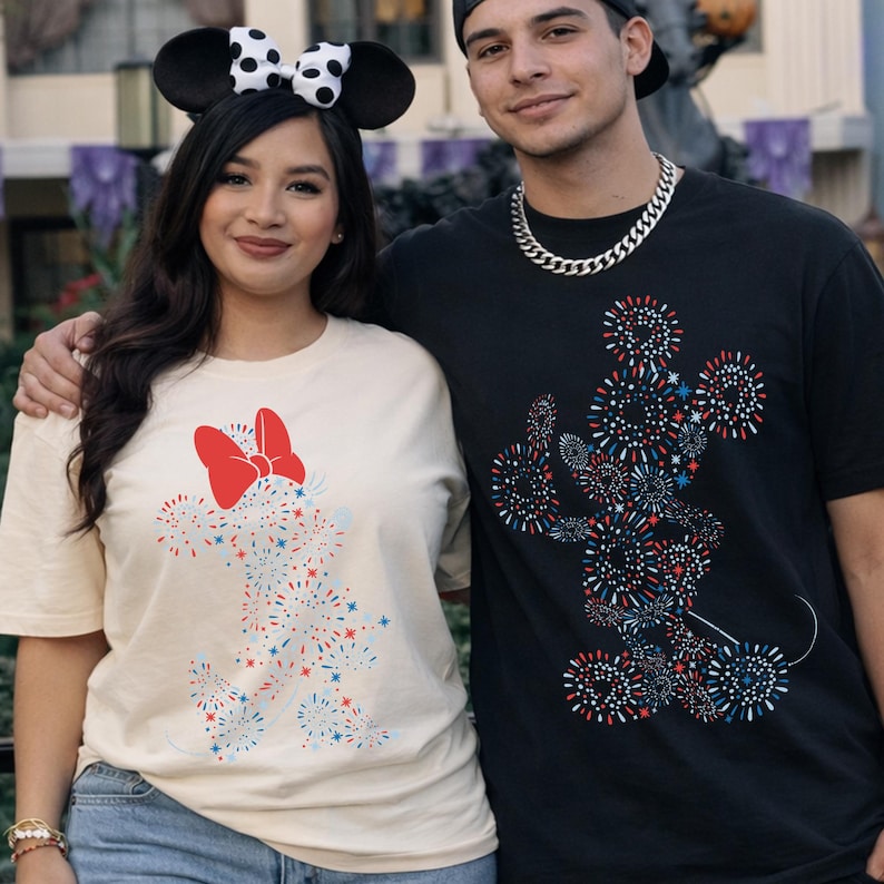 May include: Two people wearing matching t-shirts with a Mickey Mouse and Minnie Mouse design made of red, white, and blue fireworks. The woman wears a cream-colored shirt with a red bow and mouse ears. The man wears a black shirt.