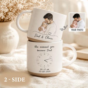 Peut inclure: Deux mugs en céramique blanche avec une base marron. L'un des mugs présente un croquis d'un père tenant un bébé, avec le texte "Dad & Olivia". L'autre mug a un design d'horloge et le texte "the moment you became Dad" avec une date et une heure.