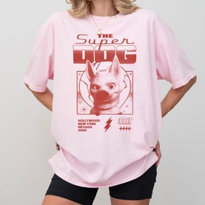 May include: Light pink t-shirt with a graphic of a dog and the text "THE SUPER DOG". The graphic is in red and features a dog's face, a lightning bolt, and the cities Hollywood, New York, Nevada, and Ohio.