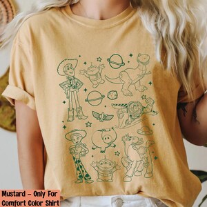 Puede incluir: Camiseta de color mostaza con dibujos lineales verdes de personajes de Toy Story. El dise&ntilde;o incluye a Woody, Buzz Lightyear, Rex y otros personajes, junto con estrellas y planetas. El texto "Mustard - Only For Comfort Color Shirt" est&aacute; en la parte inferior.