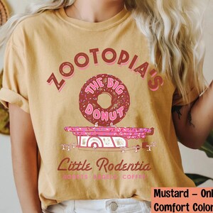 Puede incluir: Camiseta de color mostaza con un gr&aacute;fico de una rosquilla y el texto "Zootopia's The Big Donut" en rosa y marr&oacute;n. Debajo de la rosquilla hay un cami&oacute;n de comida rosa con el texto "Little Rodentia".