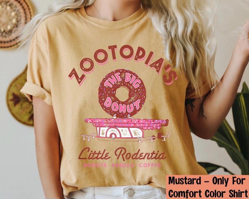 Puede incluir: Camiseta de color mostaza con un gr&aacute;fico de una rosquilla y el texto "Zootopia's The Big Donut" en rosa y marr&oacute;n. Debajo de la rosquilla hay un cami&oacute;n de comida rosa con el texto "Little Rodentia".