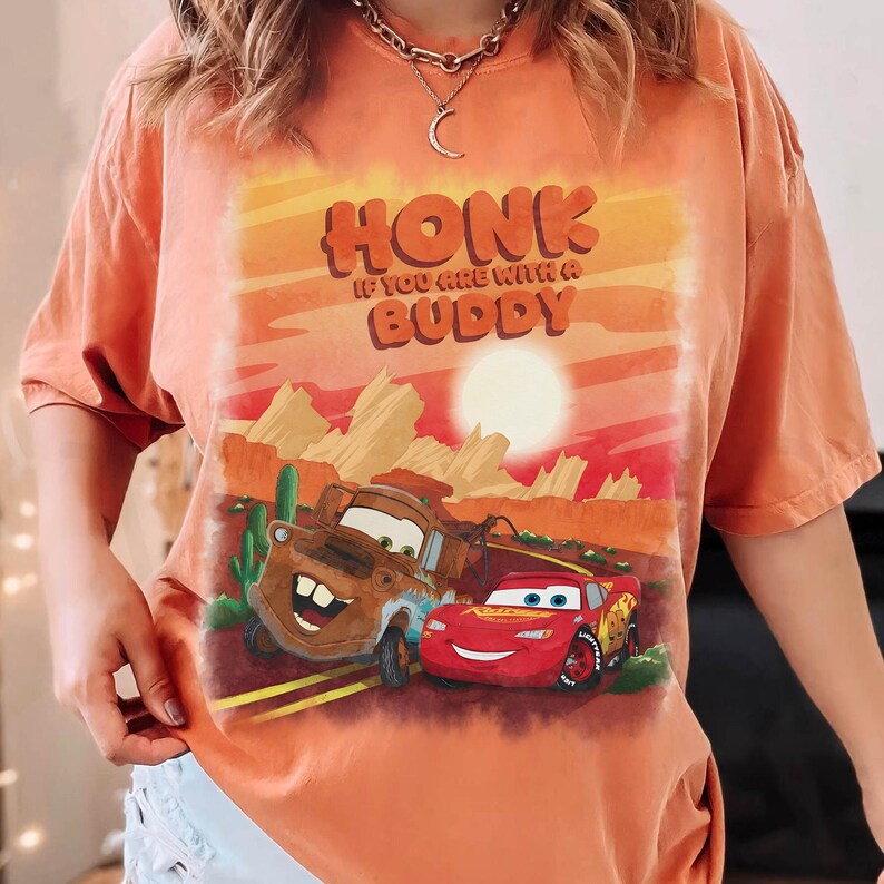 May include: Orange t-shirt featuring a graphic of the characters Mater and Lightning McQueen from the movie Cars, with the text "HONK IF YOU ARE WITH A BUDDY" in a desert landscape.