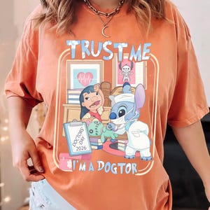 Pu&ograve; includere: T-shirt arancione con grafica raffigurante Lilo e Stitch in abito da dottore. Il design include il testo "TRUST ME" e "I'M A DOGTOR" con un cartello "DOCTORS' DAY 2026". La maglietta ha girocollo e maniche corte.