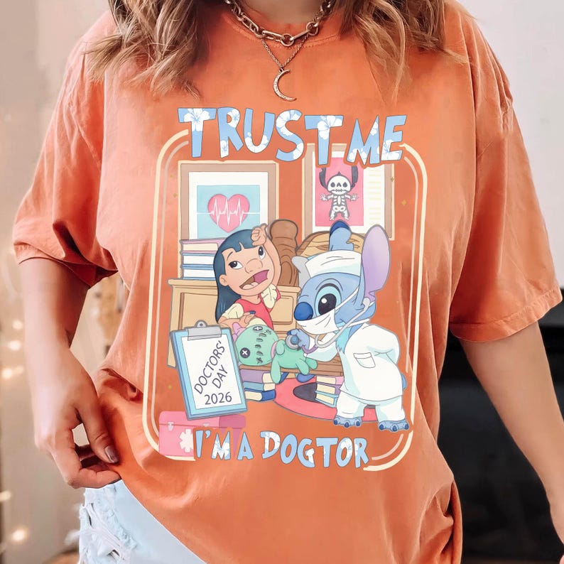 Pu&ograve; includere: T-shirt arancione con grafica raffigurante Lilo e Stitch in abito da dottore. Il design include il testo "TRUST ME" e "I'M A DOGTOR" con un cartello "DOCTORS' DAY 2026". La maglietta ha girocollo e maniche corte.