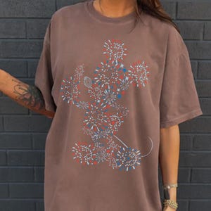 May include: Brown t-shirt featuring a stylized Mickey Mouse design composed of red, white, and blue fireworks. The graphic is centered on the front of the shirt. The shirt has short sleeves and a relaxed fit.