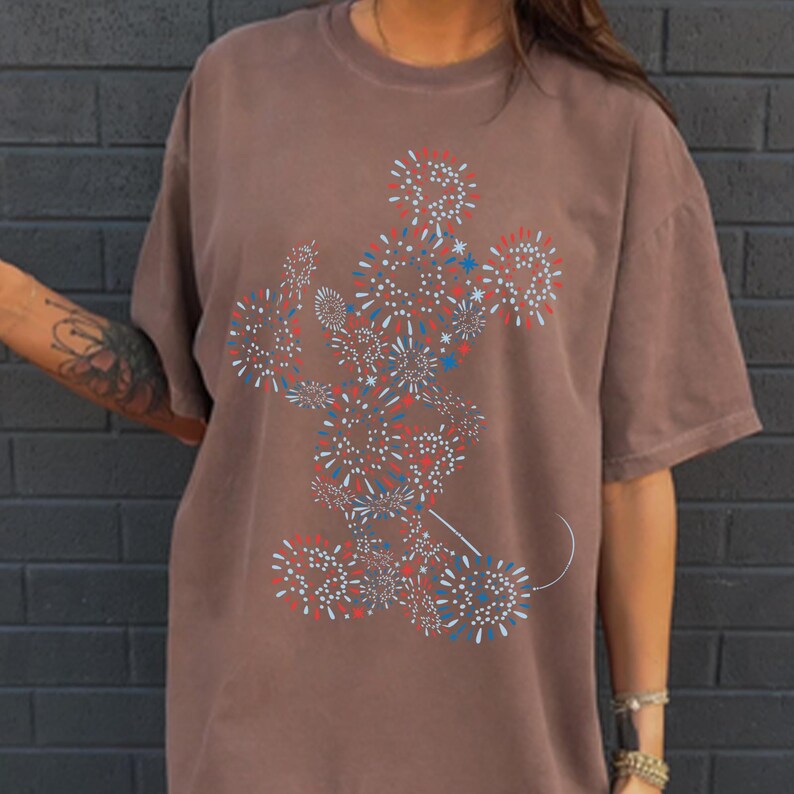 May include: Brown t-shirt featuring a stylized Mickey Mouse design composed of red, white, and blue fireworks. The graphic is centered on the front of the shirt. The shirt has short sleeves and a relaxed fit.