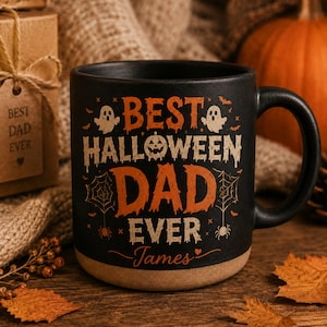 May include: A black ceramic mug with a tan base featuring orange and cream text that reads BEST HALLOWEEN DAD EVER James. The design includes illustrations of two ghosts, a jack-o'-lantern, spiderwebs, spiders, and small bats.
