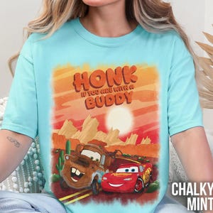 May include: A light blue t-shirt featuring a graphic of the Cars characters Mater and Lightning McQueen in a desert landscape. The text "HONK IF YOU ARE WITH A BUDDY" is printed above the cars in orange letters. The words "CHALKY MINT" are printed on the bottom right.