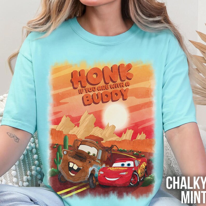 May include: A light blue t-shirt featuring a graphic of the Cars characters Mater and Lightning McQueen in a desert landscape. The text "HONK IF YOU ARE WITH A BUDDY" is printed above the cars in orange letters. The words "CHALKY MINT" are printed on the bottom right.