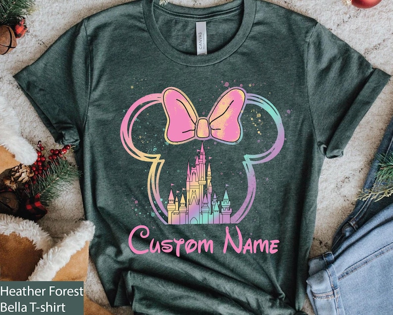 K&ouml;nnte beinhalten: Heather Forest Green Bella T-Shirt mit einem farbenfrohen Minnie Mouse-Design. Das Design umfasst eine rosa Schleife, ein Schloss und die Worte "Custom Name" in Pink. Das Design ist in Regenbogenfarben umrandet.