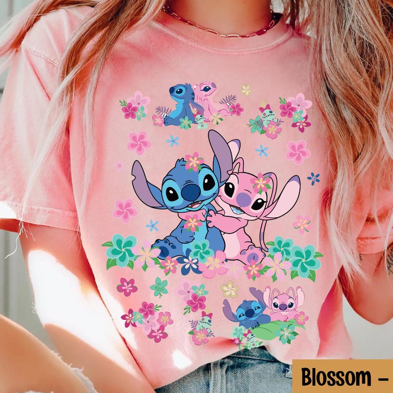 May include: Pink t-shirt featuring a graphic of Stitch and Angel embracing, surrounded by colorful flowers. The design includes Stitch and Angel in various poses, with the word "Blossom" in a rectangular box at the bottom.
