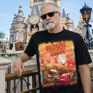 May include: Black t-shirt featuring a graphic of Mater and Lightning McQueen from the movie Cars, with the text "HONK IF YOU ARE WITH A BUDDY." The background shows a desert landscape. The image is set against a backdrop of a castle.
