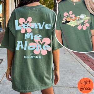 Puede incluir: Camiseta verde claro con la frase "Leave Me Alone" en azul claro, detalles de flores rosas y un dibujo de un personaje relaj&aacute;ndose con un tocadiscos. El logotipo de Disney Lilo & Stitch est&aacute; en la parte inferior. Una camiseta informal y c&oacute;moda.