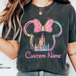 K&ouml;nnte beinhalten: Dunkelgraues T-Shirt mit einem farbenfrohen Minnie-Mouse-Design, einem Schloss und dem Text "Custom Name". Das Design umfasst eine rosa Schleife, ein Schloss und einen Regenbogen-Umriss. Das Shirt ist mit "Pepper Comfort Color" gekennzeichnet.