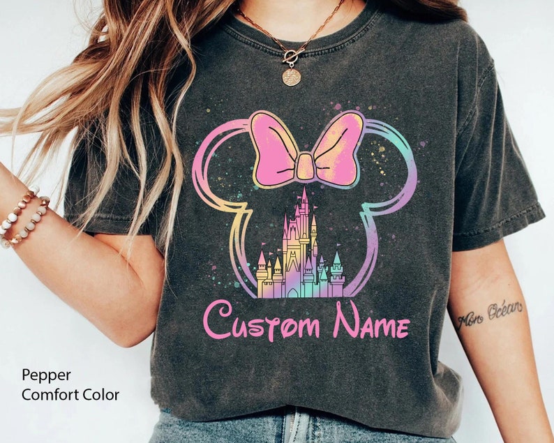 K&ouml;nnte beinhalten: Dunkelgraues T-Shirt mit einem farbenfrohen Minnie-Mouse-Design, einem Schloss und dem Text "Custom Name". Das Design umfasst eine rosa Schleife, ein Schloss und einen Regenbogen-Umriss. Das Shirt ist mit "Pepper Comfort Color" gekennzeichnet.