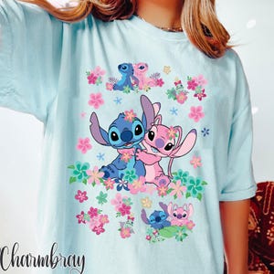 May include: Light blue t-shirt featuring a graphic of Stitch and Angel surrounded by pink and purple flowers. The design includes multiple images of the characters. The word "Charmbray" is printed in a cursive font at the bottom left.