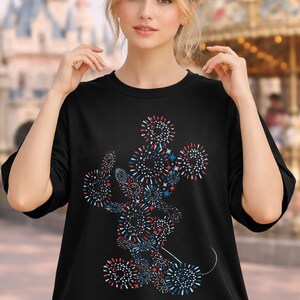 May include: Black t-shirt featuring a colorful firework design in the shape of a famous cartoon character. The design incorporates red, white, and blue elements. The shirt is a crew neck style.