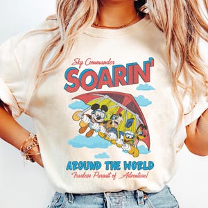 Puede incluir: Una camiseta blanca con un gr&aacute;fico de estilo vintage que presenta a Mickey Mouse, Pluto y Goofy volando en un ala delta. El texto en la camiseta dice "Sky Commander Soarin' Around the World Fearless Pursuit of Adventure!"
