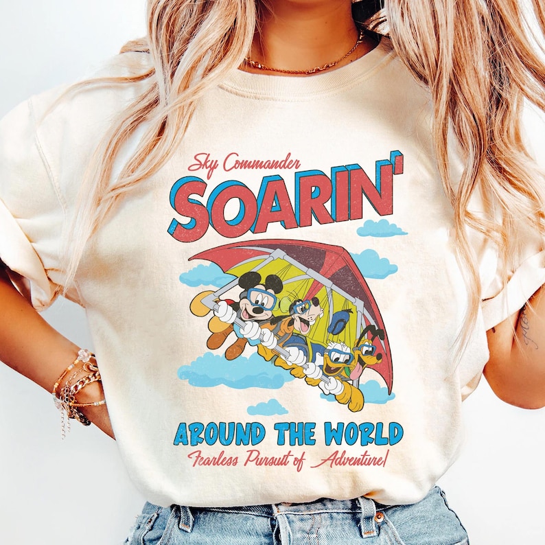 Puede incluir: Una camiseta blanca con un gr&aacute;fico de estilo vintage que presenta a Mickey Mouse, Pluto y Goofy volando en un ala delta. El texto en la camiseta dice "Sky Commander Soarin' Around the World Fearless Pursuit of Adventure!"
