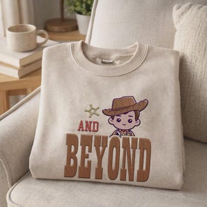 Puede incluir: Sudadera beige con un personaje de vaquero de dibujos animados bordado con un sombrero marr&oacute;n y las palabras "AND BEYOND" en marr&oacute;n. La sudadera est&aacute; doblada sobre un sill&oacute;n beige.