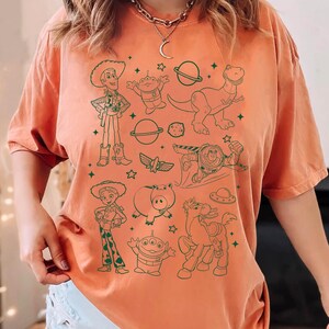 Puede incluir: Camiseta naranja con un dise&ntilde;o en l&iacute;nea verde de personajes de dibujos animados, incluyendo un vaquero, extraterrestres, un dinosaurio y un guardabosques espacial. El dise&ntilde;o tambi&eacute;n incluye planetas, estrellas y un cerdo. La camiseta tiene mangas cortas y cuello redondo.