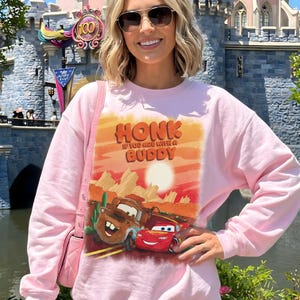 May include: Pink sweatshirt featuring a graphic of Mater and Lightning McQueen from the movie Cars, with the text "HONK IF YOU ARE WITH A BUDDY". The sweatshirt is a crew neck style.