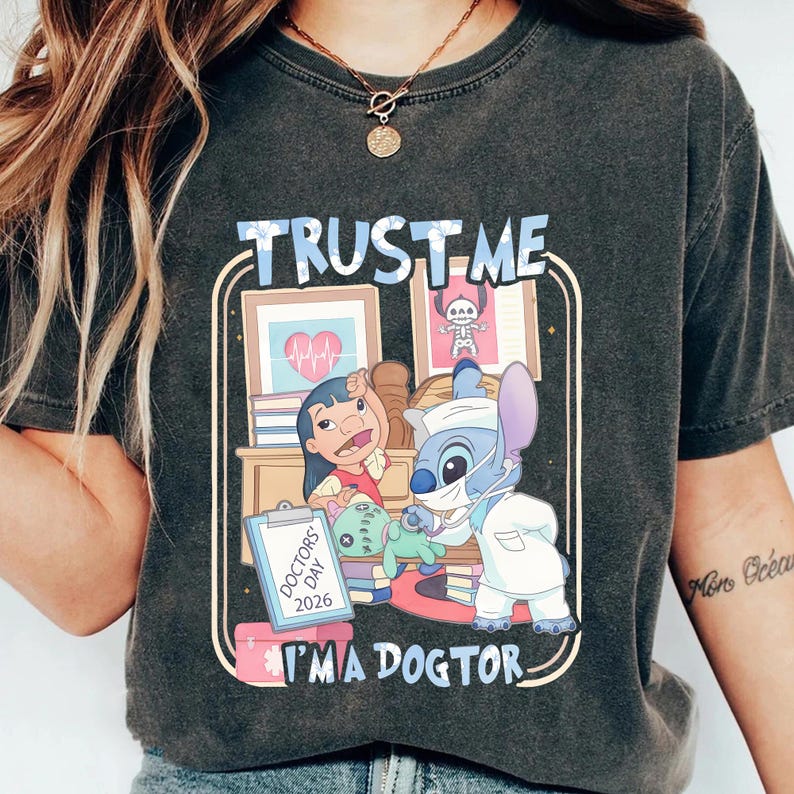 Pu&ograve; includere: T-shirt grigio scuro con una grafica a cartoni animati con la scritta "TRUST ME I'M A DOCTOR". La grafica include i personaggi di Lilo e Stitch in un ambiente medico, con attrezzature mediche e la scritta "DOCTORS DAY 2026".