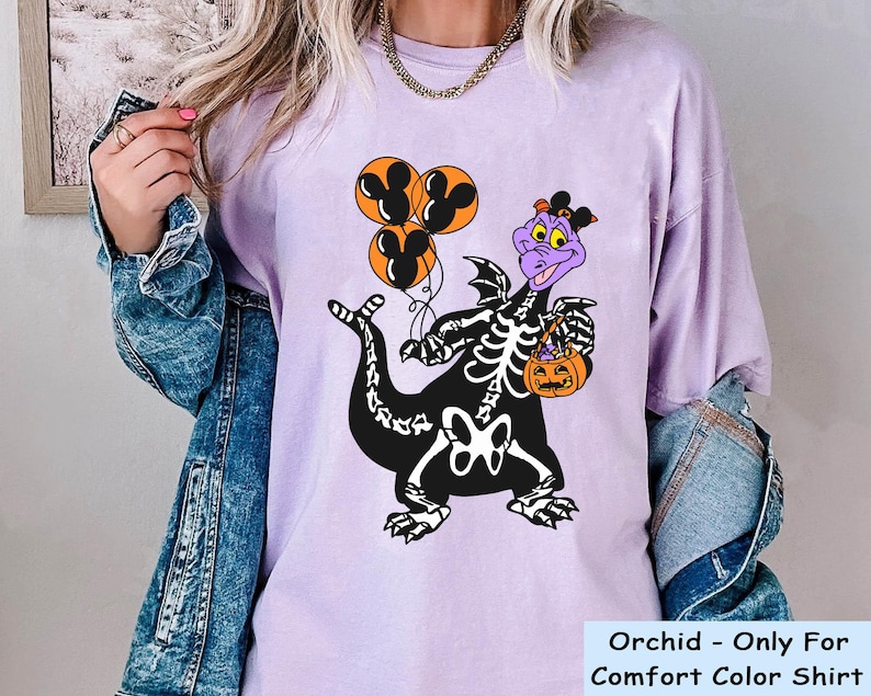 May include: An orchid-coloured t-shirt featuring a graphic of a purple dragon wearing a skeleton costume, holding three orange Mickey Mouse-shaped balloons and an orange jack-o'-lantern bucket. The shirt is layered under a blue denim jacket.